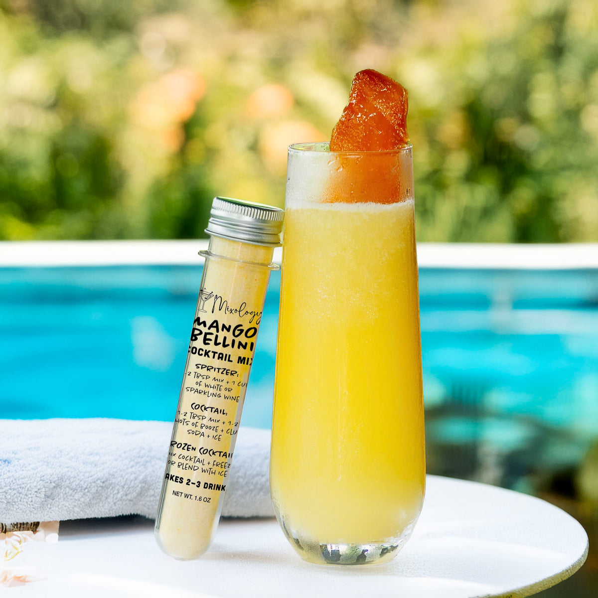 A tall clear glass filled with a bright yellow-orange cocktail sits on a white table beside a small labeled tube of mango melon cocktail mix, with a blurred pool and greenery in the background.