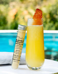 A tall clear glass filled with a bright yellow-orange cocktail sits on a white table beside a small labeled tube of mango melon cocktail mix, with a blurred pool and greenery in the background.