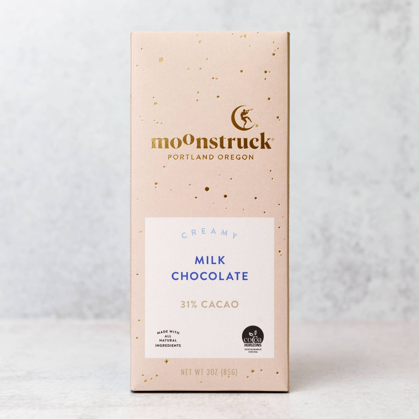 Moonstruck creamy chocolate bar in its rectangular-shaped package that is pink with gold spots, gold lettering, and a white label with blue letters and two symbols in black on a marble-like background. 
