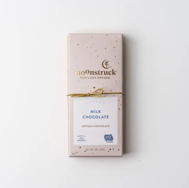 Moonstruck creamy chocolate bar in its rectangular-shaped package that is pink with gold spots, gold lettering, and a white label with blue letters and two symbols in blue wrapped in a gold bow on a white background. 