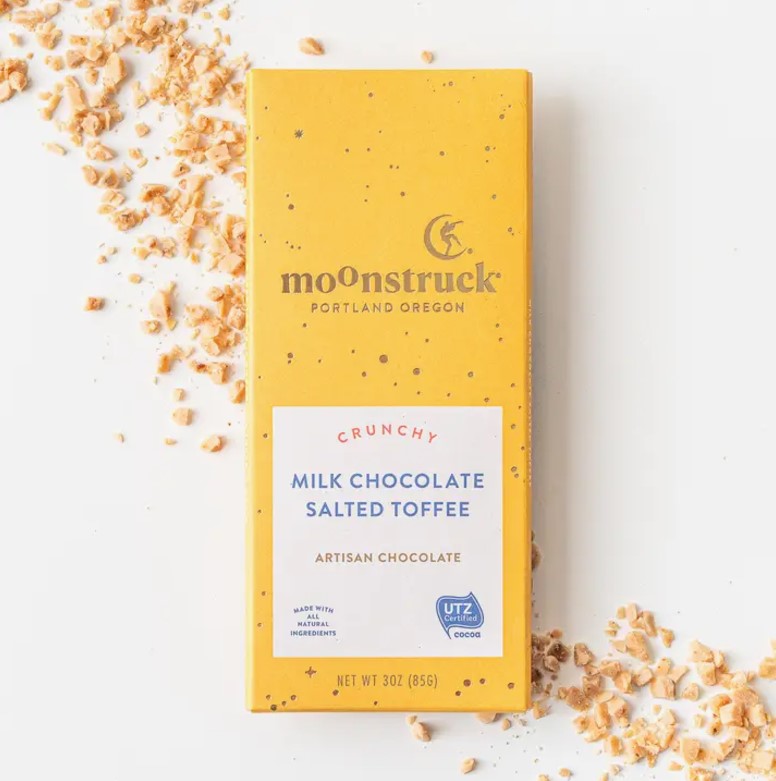 Moonstruck milk chocolate salted toffee bar is featured in its yellow rectangular package with gold speckles and gold lettering title. With a white imprinted label with blue, dark pink and light brown lettering information and details.  Pieces of almonds around it on a white background. 
