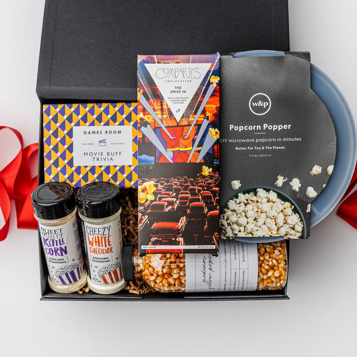 The movie night gift box is featured. A popcorn popper, a box of movie trivia, a bottle of popcorn kernels, two popcorn seasonings, and a  Compartés drive-in popcorn chocolate bar are arranged inside a black gift box with brown shred, with a red ribbon draped around the base. The items are neatly placed, taller elements like the popcorn popper and popcorn bottle in the back, while the trivia box sits centrally, and the seasonings are positioned in front. On a white background.  