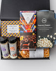 The movie night gift box is featured. A popcorn popper, a box of movie trivia, a bottle of popcorn kernels, two popcorn seasonings, and a  Compartés drive-in popcorn chocolate bar are arranged inside a black gift box with brown shred, with a red ribbon draped around the base. The items are neatly placed, taller elements like the popcorn popper and popcorn bottle in the back, while the trivia box sits centrally, and the seasonings are positioned in front. On a white background.  