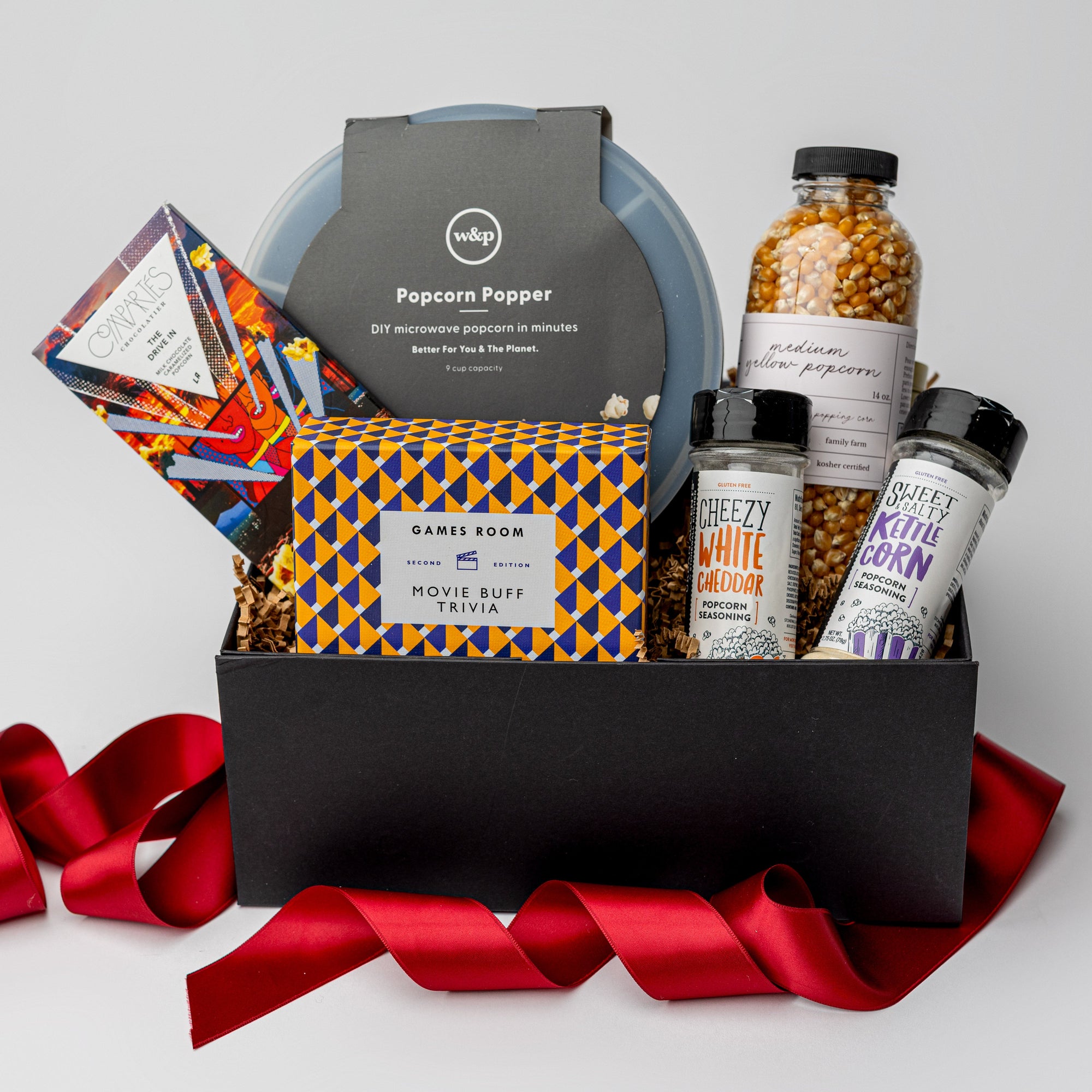 The movie night gift box is featured. A popcorn popper, a box of movie trivia, a bottle of popcorn kernels, two popcorn seasonings, and a  Compartés drive-in popcorn chocolate bar are arranged inside a black gift box with brown shred, with a red ribbon draped around the base. The items are neatly placed, taller elements like the popcorn popper and popcorn bottle in the back, while the trivia box sits centrally, and the seasonings are positioned in front. On a white background.  