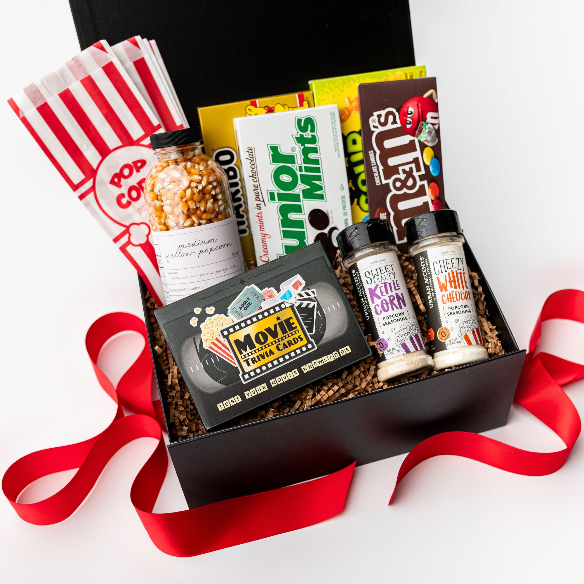A top view of the Movie Night Gift Box presented in a black gift box filled with brown shred and tied with a red ribbon, featuring a Movie Trivia Game, small batch popcorn kernels, four popcorn bags, a set of four theater box candies, and a set of popcorn seasonings in white cheddar and kettle corn flavors, all arranged on a white background.