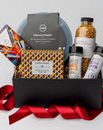 The movie night gift box is featured. A popcorn popper, a box of movie trivia, a bottle of popcorn kernels, two popcorn seasonings, and a  Compartés drive-in popcorn chocolate bar are arranged inside a black gift box with brown shred, with a red ribbon draped around the base. The items are neatly placed, taller elements like the popcorn popper and popcorn bottle in the back, while the trivia box sits centrally, and the seasonings are positioned in front. On a white background.  