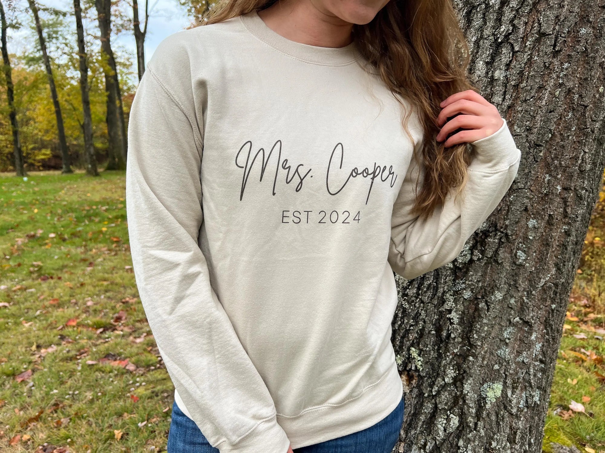 Mrs. established a custom sweatshirt in sand color in black lettering personalized with Mrs. Cooper Est. 2024, worn by a bride in wood -wood-nature-like like environment. 