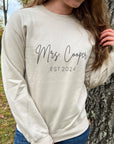 Mrs. established a custom sweatshirt in sand color in black lettering personalized with Mrs. Cooper Est. 2024, worn by a bride in wood -wood-nature-like like environment. 