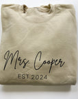 Mrs. established custom sweatshirt in sand color in black lettering personalized with Mrs. Cooper Est. 2024 in a white background.
