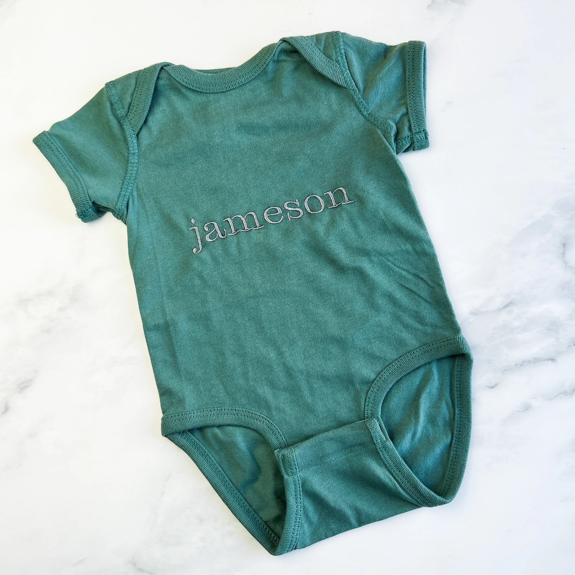Name embroidered onesie featured in basil color personalized in champagne thread with the name Jameson on a marble like background.