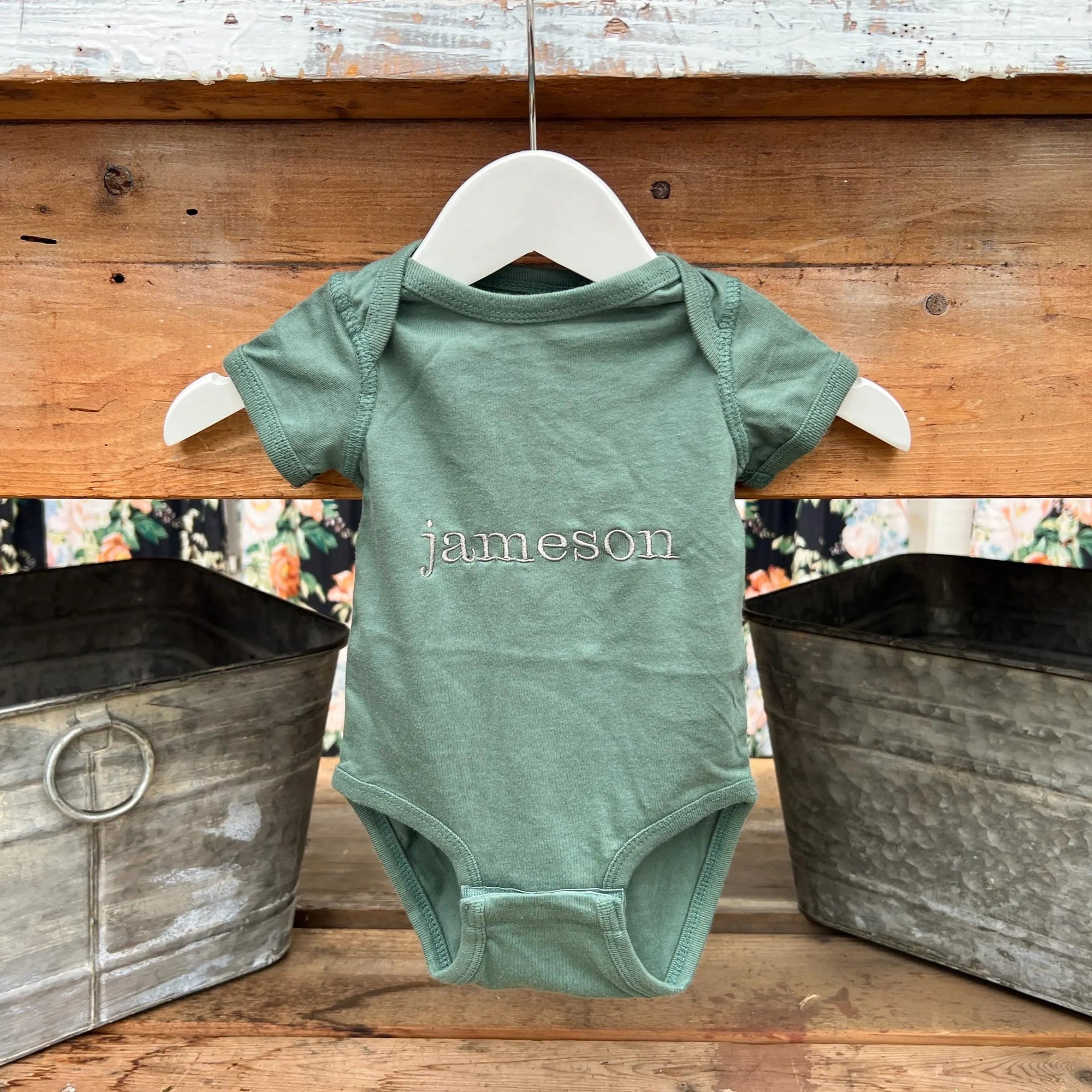 Name embroidered onesie featured in basil color, personalized in champagne thread with the name Jameson on a wooden background with two silver buckets on each side of it. 