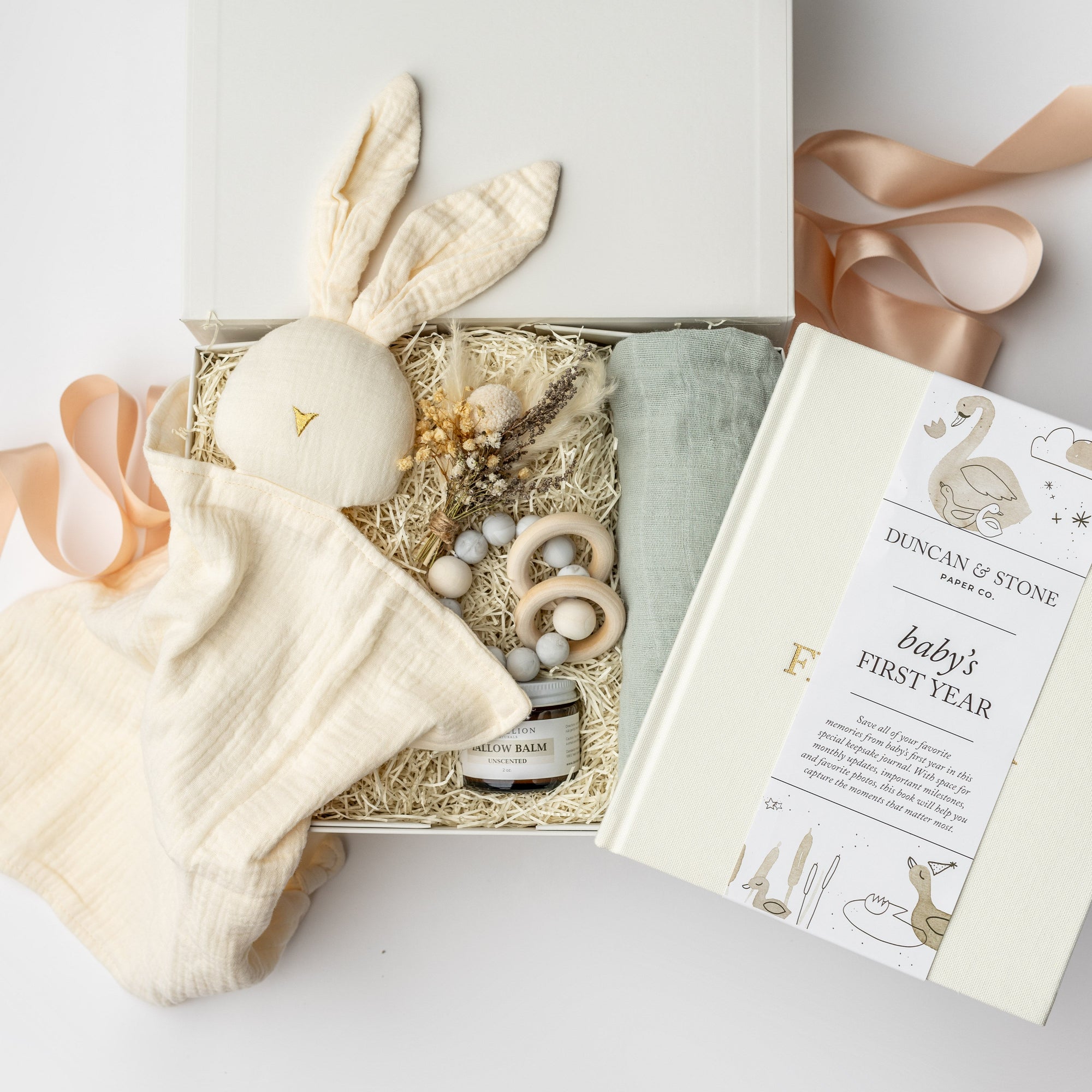 A top view of a New Baby Gift Box featuring a Baby’s First Year book, a wooden teether, a soft bunny lovey, Dandelion natural tallow balm, a handcrafted natural floral bundle, and a sage green swaddle, all arranged in a white gift box with ivory shred and tied with a blush ribbon on a white background.