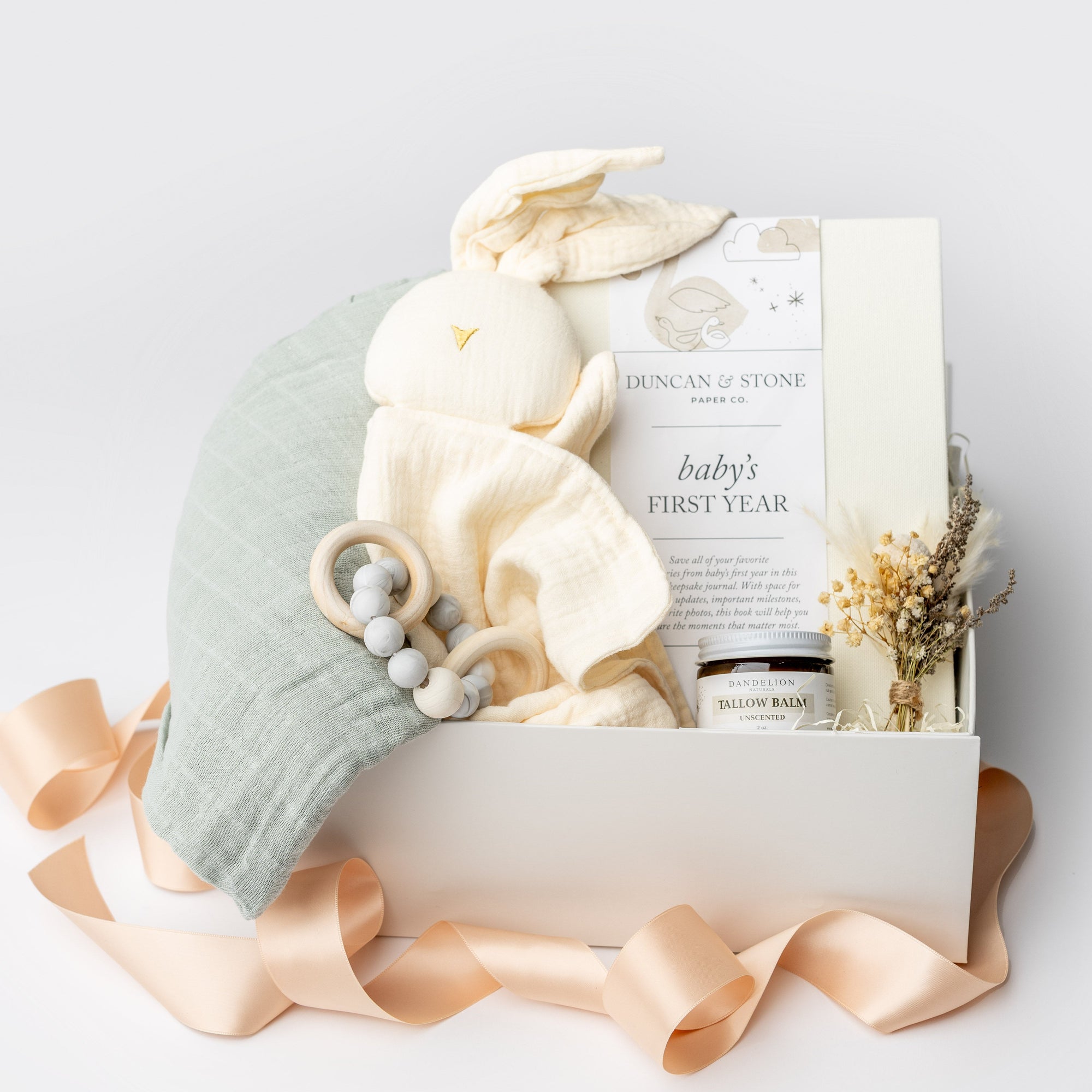 A front view of the New Baby Gift Box featuring a Baby’s First Year book, a wooden teether, a soft bunny lovey, Dandelion natural tallow balm, a handcrafted natural floral bundle, and a sage green swaddle, all arranged in a white gift box with ivory shred and tied with a blush ribbon on a white background.