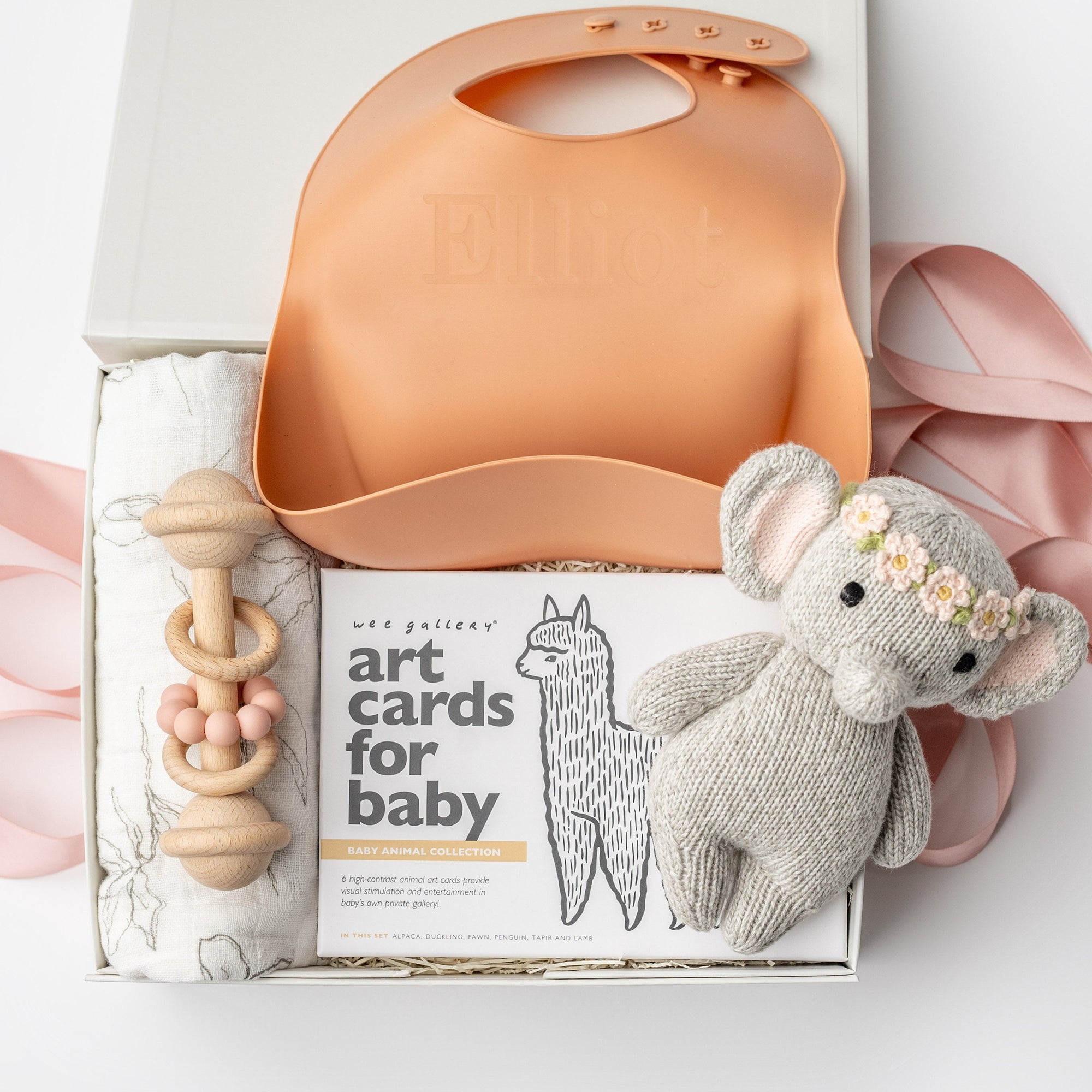 New baby girl gift box, A top view of a Baby Girl Gift Box featuring a knitted cute elephant holding roses, a floral swaddle, a wooden rattle, the book Guess How Much I Love You, and a pink silicone bib with an engraved name, all arranged in a white gift box with ivory shred and tied with a pink ribbon on a white background.