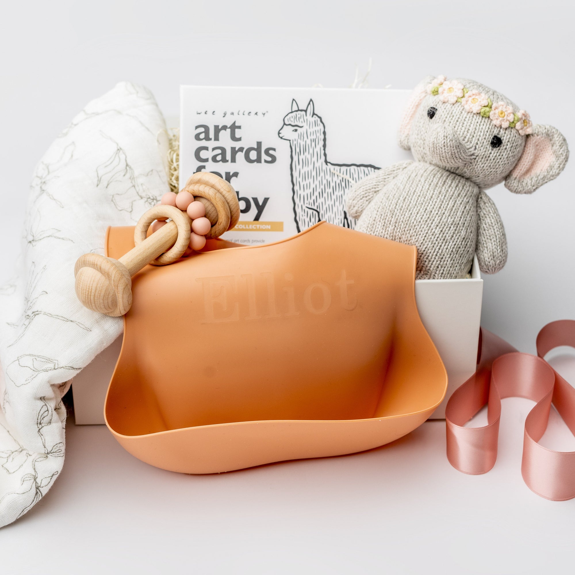 New baby girl gift box, A front view of a Baby Girl Gift Box featuring a knitted cute elephant holding roses, a floral swaddle, a wooden rattle, the book Guess How Much I Love You, and a pink silicone bib with an engraved name, all arranged in a white gift box with ivory shred and tied with a pink ribbon on a white background.