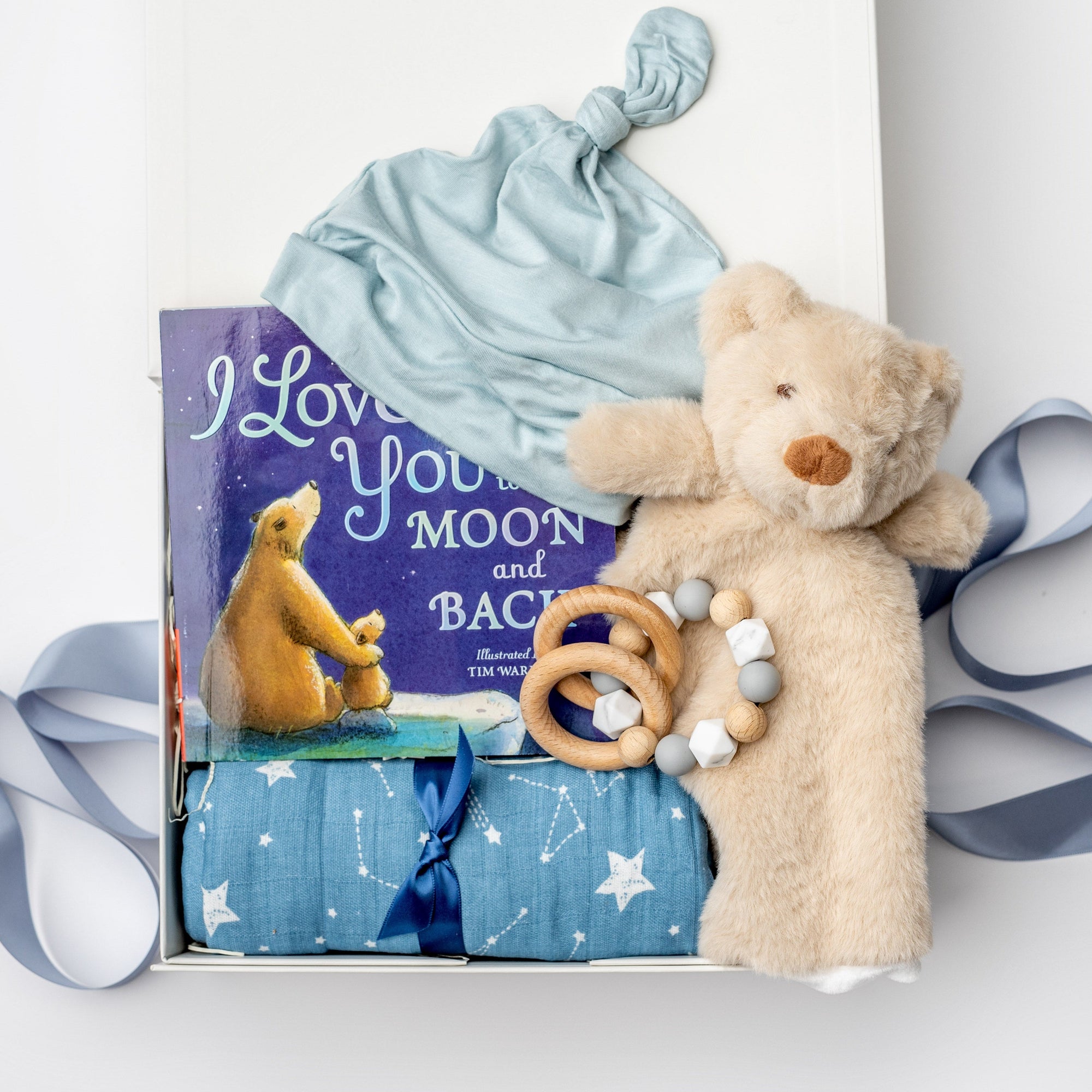 A white gift box on a white background, filled with light ivory paper shred and tied with a soft blue ribbon. Inside the box: a plush teddy bear lovey (with optional personalization), a muslin cotton swaddle blanket printed with white stars on a dusty-blue background, a soft blue stretch baby top-knot hat, a silicone and wood baby teether consisting of two wooden rings and a grey-and-white bead loop, and a board book titled “I Love You to the Moon and Back.”