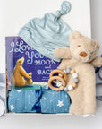 A white gift box on a white background, filled with light ivory paper shred and tied with a soft blue ribbon. Inside the box: a plush teddy bear lovey (with optional personalization), a muslin cotton swaddle blanket printed with white stars on a dusty-blue background, a soft blue stretch baby top-knot hat, a silicone and wood baby teether consisting of two wooden rings and a grey-and-white bead loop, and a board book titled “I Love You to the Moon and Back.”