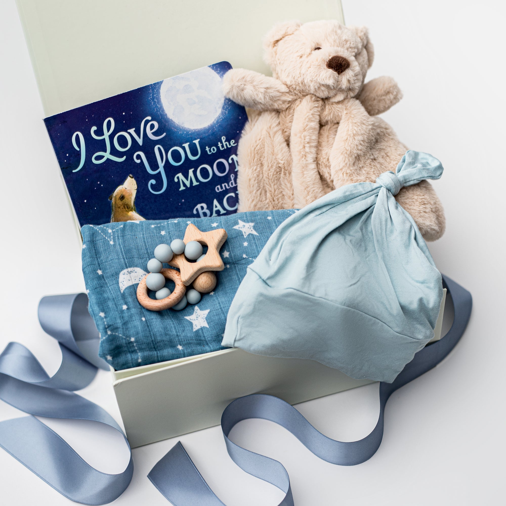 A white gift box on a white background, filled with light ivory paper shred and tied with a soft blue ribbon. Inside the box: a plush teddy bear lovey (with optional personalization), a muslin cotton swaddle blanket printed with white stars on a dusty-blue background, a soft blue stretch baby top-knot hat, a silicone and wood baby teether consisting of two wooden rings and a grey-and-white bead loop, and a board book titled “I Love You to the Moon and Back.”