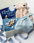 A white gift box on a white background, filled with light ivory paper shred and tied with a soft blue ribbon. Inside the box: a plush teddy bear lovey (with optional personalization), a muslin cotton swaddle blanket printed with white stars on a dusty-blue background, a soft blue stretch baby top-knot hat, a silicone and wood baby teether consisting of two wooden rings and a grey-and-white bead loop, and a board book titled “I Love You to the Moon and Back.”