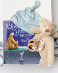 A white gift box on a white background, filled with light ivory paper shred and tied with a soft blue ribbon. Inside the box: a plush teddy bear lovey (with optional personalization), a muslin cotton swaddle blanket printed with white stars on a dusty-blue background, a soft blue stretch baby top-knot hat, a silicone and wood baby teether consisting of two wooden rings and a grey-and-white bead loop, and a board book titled “I Love You to the Moon and Back.”