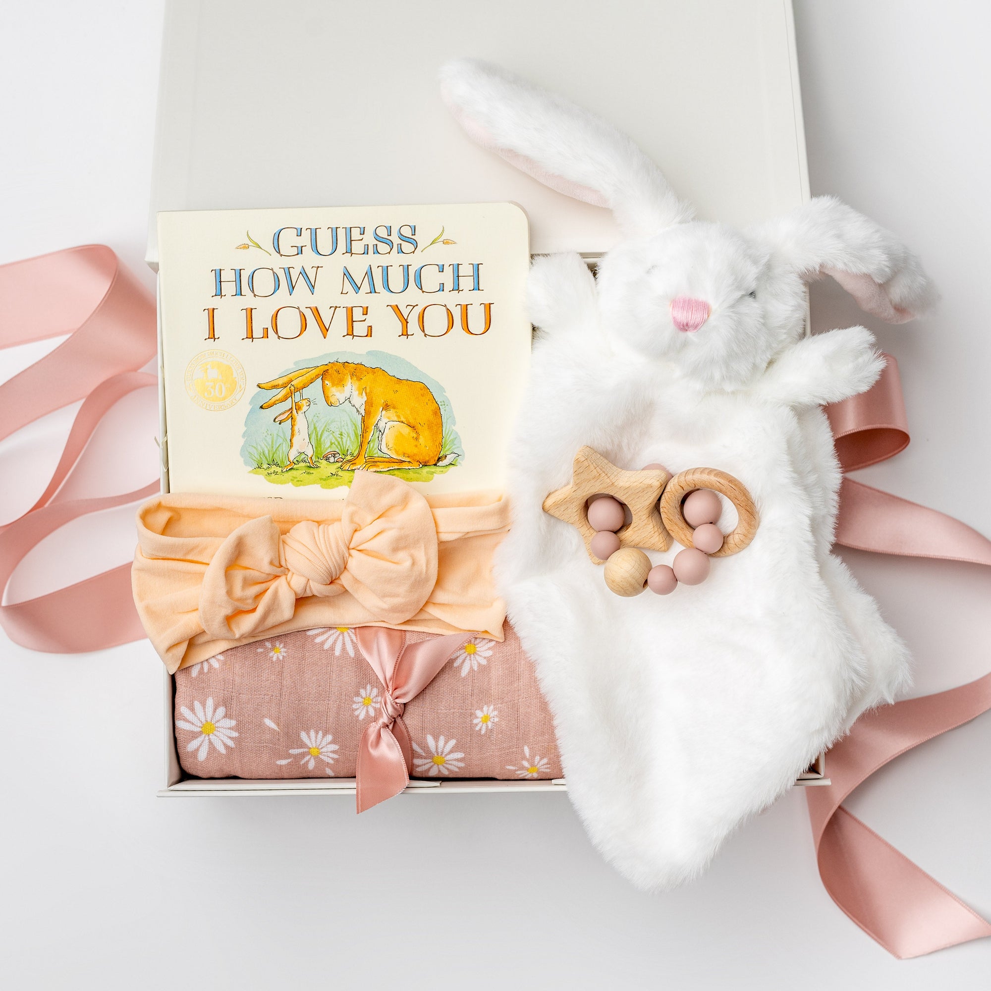 Welcome baby girl gift box, A white gift box filled with ivory paper shred and tied with a soft pink ribbon on a white background. Inside the box is a plush white bunny lovey (optionally embroidered), a cotton muslin swaddle blanket in a pink floral print, a peach-colored stretch bow headband, a silicone and wood baby teether shaped with beads and a star, and a board book titled “Guess How Much I Love You.”
