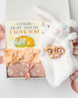 Welcome baby girl gift box, A white gift box filled with ivory paper shred and tied with a soft pink ribbon on a white background. Inside the box is a plush white bunny lovey (optionally embroidered), a cotton muslin swaddle blanket in a pink floral print, a peach-colored stretch bow headband, a silicone and wood baby teether shaped with beads and a star, and a board book titled “Guess How Much I Love You.”