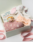 Welcome baby girl gift box, A white gift box filled with ivory paper shred and tied with a soft pink ribbon on a white background. Inside the box is a plush white bunny lovey (optionally embroidered), a cotton muslin swaddle blanket in a pink floral print, a peach-colored stretch bow headband, a silicone and wood baby teether shaped with beads and a star, and a board book titled “Guess How Much I Love You.”