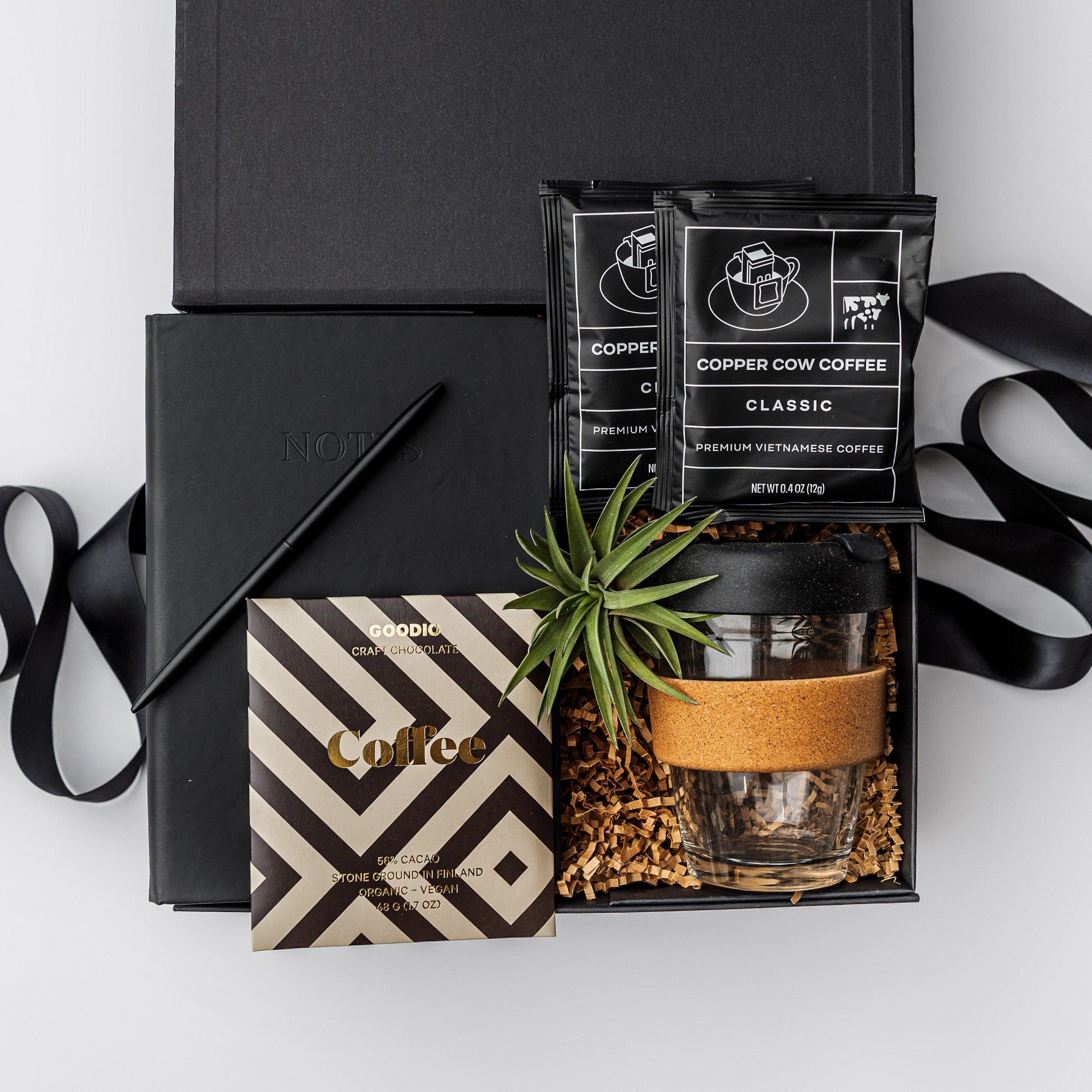 New Hire Employee Onboarding Gift Box, A top view of a modern coffee-themed gift set presented in a black gift box filled with brown shred and tied with a black ribbon, featuring a black softcover journal, a minimalist black bullet pen, a 12 oz. glass travel mug with a cork sleeve, two Copper Cow artisan pour-over coffee packets, a Goodio coffee chocolate bar (56% cacao), and a small live Tillandsia air plant.