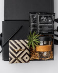 New Hire Employee Onboarding Gift Box, A top view of a modern coffee-themed gift set presented in a black gift box filled with brown shred and tied with a black ribbon, featuring a black softcover journal, a minimalist black bullet pen, a 12 oz. glass travel mug with a cork sleeve, two Copper Cow artisan pour-over coffee packets, a Goodio coffee chocolate bar (56% cacao), and a small live Tillandsia air plant.