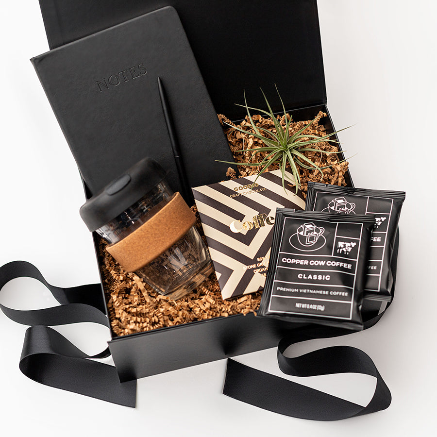 New Hire Employee Onboarding Gift Box, A top view of a modern coffee-themed gift set presented in a black gift box filled with brown shred and tied with a black ribbon, featuring a black softcover journal, a minimalist black bullet pen, a 12 oz. glass travel mug with a cork sleeve, two Copper Cow artisan pour-over coffee packets, a Goodio coffee chocolate bar (56% cacao), and a small live Tillandsia air plant.