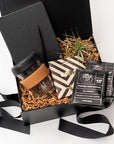 New Hire Employee Onboarding Gift Box, A top view of a modern coffee-themed gift set presented in a black gift box filled with brown shred and tied with a black ribbon, featuring a black softcover journal, a minimalist black bullet pen, a 12 oz. glass travel mug with a cork sleeve, two Copper Cow artisan pour-over coffee packets, a Goodio coffee chocolate bar (56% cacao), and a small live Tillandsia air plant.