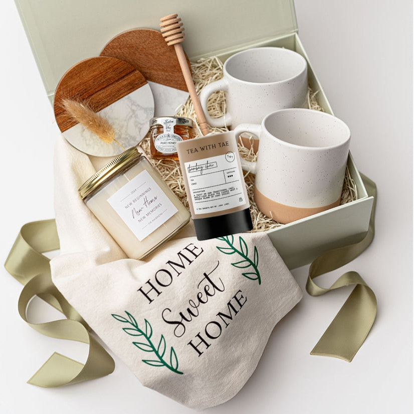 A top view of the New Home Housewarming Gift Box featuring a set of two marble and wood coasters, a “Home Sweet Home” tea towel, a 9 oz. hand-poured candle labeled “New Beginnings, New Home, New Memories” (optionally personalized with a family name), a mini tea tube of jasmine green tea, a mini jar of honey with a wooden honey drizzler, and a set of two speckled stoneware mugs, all arranged in a white gift box with ivory shred and tied with a green ribbon on a white background.