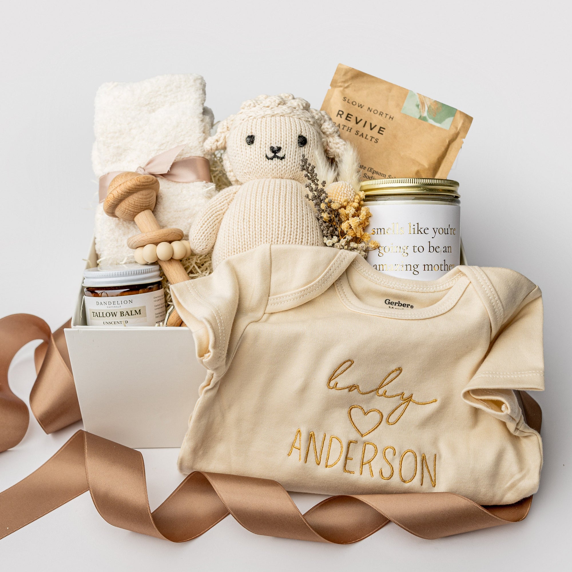 A white gift box with ivory paper shred and a brown ribbon on a white background. Inside the box are an 8-oz hand-poured lavender soy candle labeled “Amazing Mother,” a pair of cozy cloud plush socks, a Gerber onesie with personalized embroidery, Dandelion Naturals tallow balm, a Cuddle + Kind knit lamb, a wooden rattle, Slow North “Revive” natural bath salts, and a handcrafted dried floral mini bouquet — a “New Mom & Baby” gift box.
