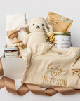 A white gift box with ivory paper shred and a brown ribbon on a white background. Inside the box are an 8-oz hand-poured lavender soy candle labeled “Amazing Mother,” a pair of cozy cloud plush socks, a Gerber onesie with personalized embroidery, Dandelion Naturals tallow balm, a Cuddle + Kind knit lamb, a wooden rattle, Slow North “Revive” natural bath salts, and a handcrafted dried floral mini bouquet — a “New Mom & Baby” gift box.