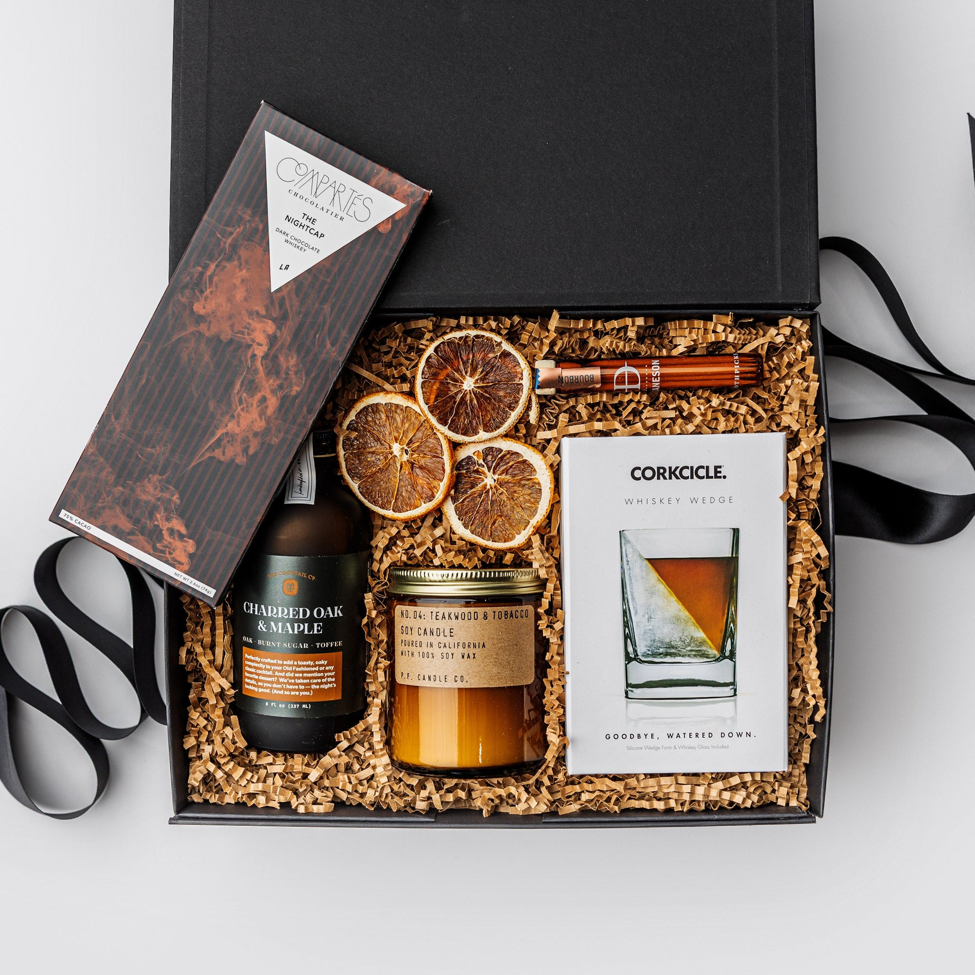 Nightcap Whiskey Cocktail Curated Gift Box, A top view of the Night Cap Gift Box presented in a black gift box filled with brown shred and tied with a black ribbon, featuring a Corkcicle whiskey glass with ice wedge, a Compartes Nightcap dark chocolate bar, scotch-infused wooden toothpicks, a charred oak & maple syrup artisan cocktail mix, a PF Candle Co. 7.2 oz Teakwood & Tobacco candle, and a crispy orange slices snack pack, all arranged on a white background.