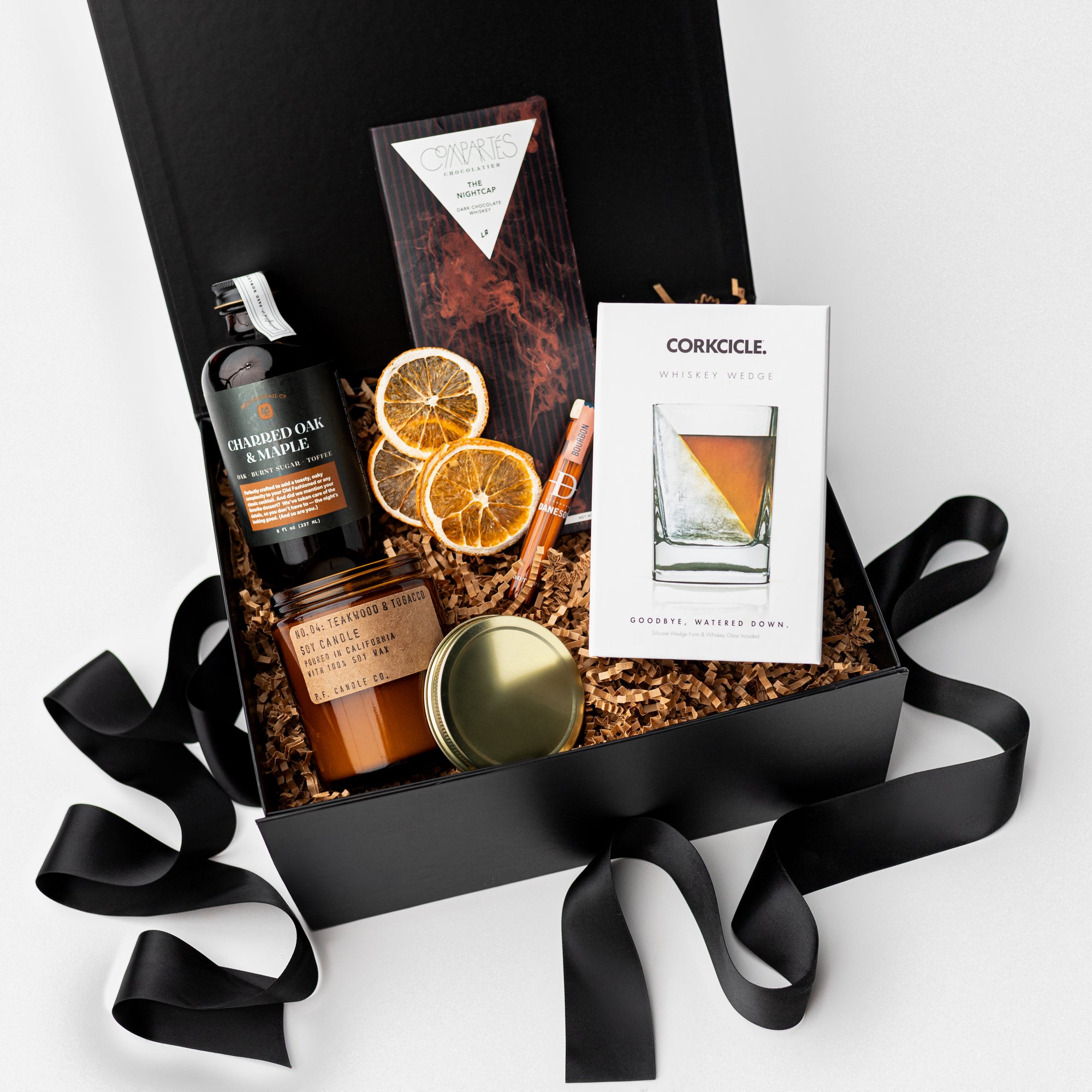 Nightcap Whiskey Cocktail Curated Gift Box, A top view of the Night Cap Gift Box presented in a black gift box filled with brown shred and tied with a black ribbon, featuring a Corkcicle whiskey glass with ice wedge, a Compartes Nightcap dark chocolate bar, scotch-infused wooden toothpicks, a charred oak & maple syrup artisan cocktail mix, a PF Candle Co. 7.2 oz Teakwood & Tobacco candle, and a crispy orange slices snack pack, all arranged on a white background.