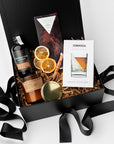 Nightcap Whiskey Cocktail Curated Gift Box, A top view of the Night Cap Gift Box presented in a black gift box filled with brown shred and tied with a black ribbon, featuring a Corkcicle whiskey glass with ice wedge, a Compartes Nightcap dark chocolate bar, scotch-infused wooden toothpicks, a charred oak & maple syrup artisan cocktail mix, a PF Candle Co. 7.2 oz Teakwood & Tobacco candle, and a crispy orange slices snack pack, all arranged on a white background.