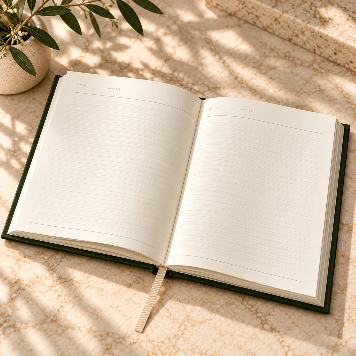 An open lined notebook or journal with a dark green cover lies on a light stone surface in soft dappled sunlight, with a plant leaf in the upper left corner.