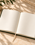 An open lined notebook or journal with a dark green cover lies on a light stone surface in soft dappled sunlight, with a plant leaf in the upper left corner.