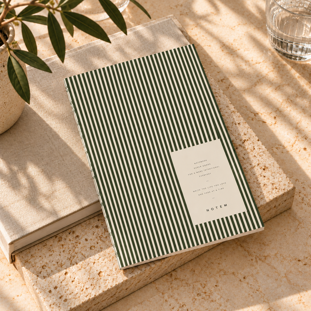 A notebook with dark green and white vertical stripes and a white label reading 'NOTEN', styled on a beige stone block with a plant and glass in a sunlit setting.