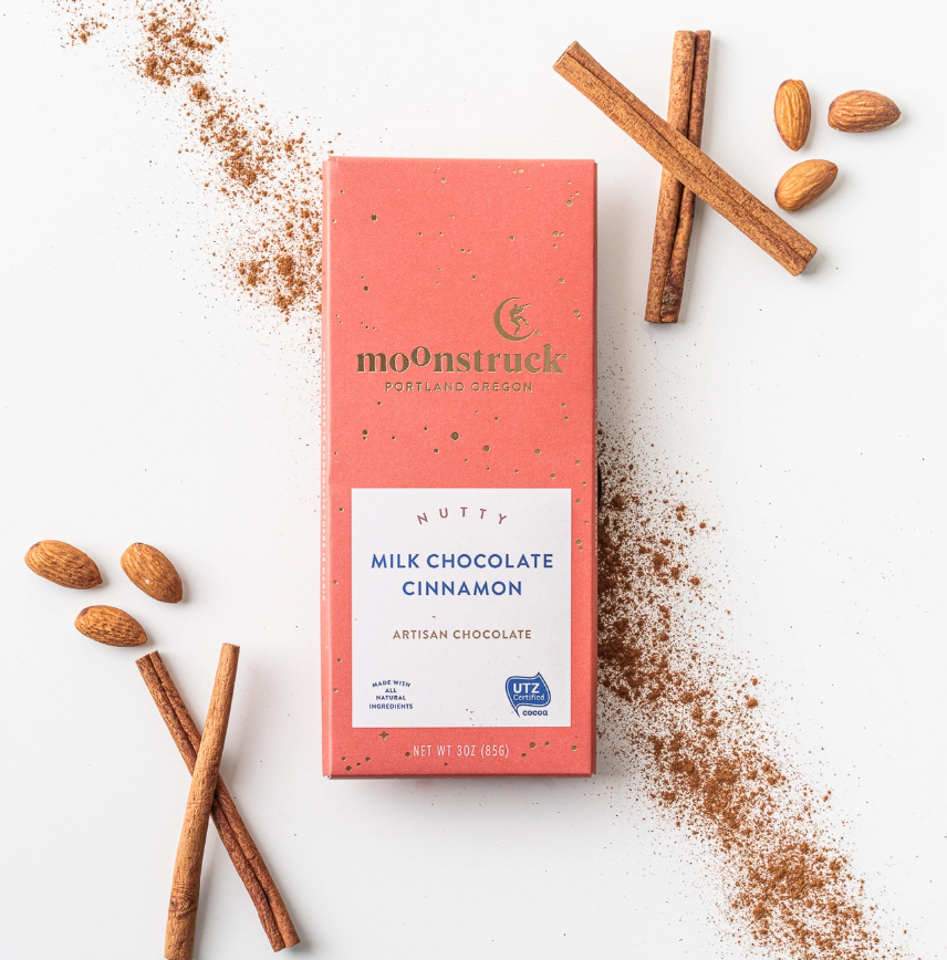 Nutty milk chocolate cinnamon bar is featured in its pink rectangular-shaped package that has gold specks, gold lettering and a label in white printed on it using brown and blue lettering. Cinnamon powder, sticks and six almonds around it on a white background. 