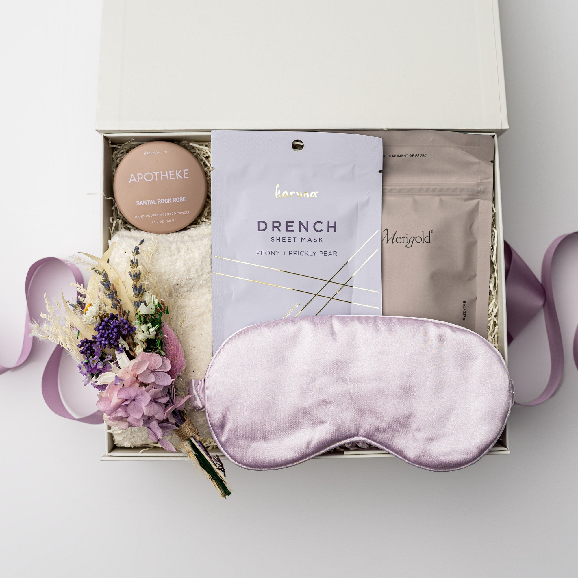 A top view of a relaxing self-care gift set presented in a white gift box filled with ivory shred and tied with a lavender ribbon, featuring a satin sleep mask, a 2 oz. Santal Rock Rose candle, a Drench sleep mask, an 8 oz. lavender oat bath soak, cloud socks, and a handmade dried floral mini bouquet, all arranged on a white background.