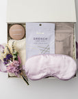 A top view of a relaxing self-care gift set presented in a white gift box filled with ivory shred and tied with a lavender ribbon, featuring a satin sleep mask, a 2 oz. Santal Rock Rose candle, a Drench sleep mask, an 8 oz. lavender oat bath soak, cloud socks, and a handmade dried floral mini bouquet, all arranged on a white background.