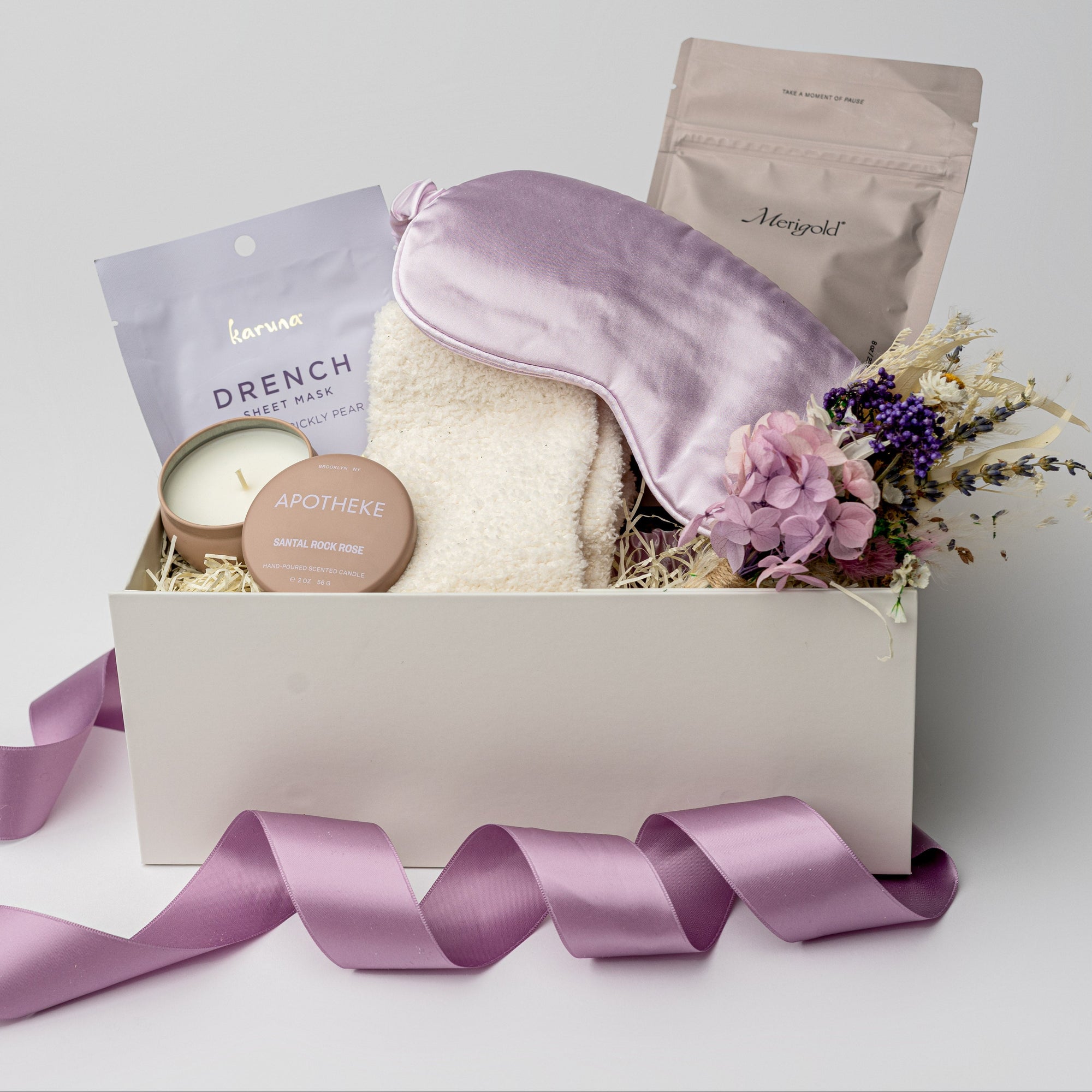 A front view of a relaxing self-care gift set presented in a white gift box filled with ivory shred and tied with a lavender ribbon, featuring a satin sleep mask, a 2 oz. Santal Rock Rose candle, a Drench sleep mask, an 8 oz. lavender oat bath soak, cloud socks, and a handmade dried floral mini bouquet, all arranged on a white background.