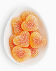Sugarfina Peach Bellini Gummies in a white tray, showing peach-colored gummy candies inside, each shaped like miniature heart. On a white background. 
