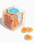 Sugarfina Peach Bellini Gummies in a clear plastic cube-shaped package, showing peach-colored gummy candies inside, each shaped like miniature hearts, with the Sugarfina logo and 'Peach Bellini' label on the front. Three outside on a white background. 