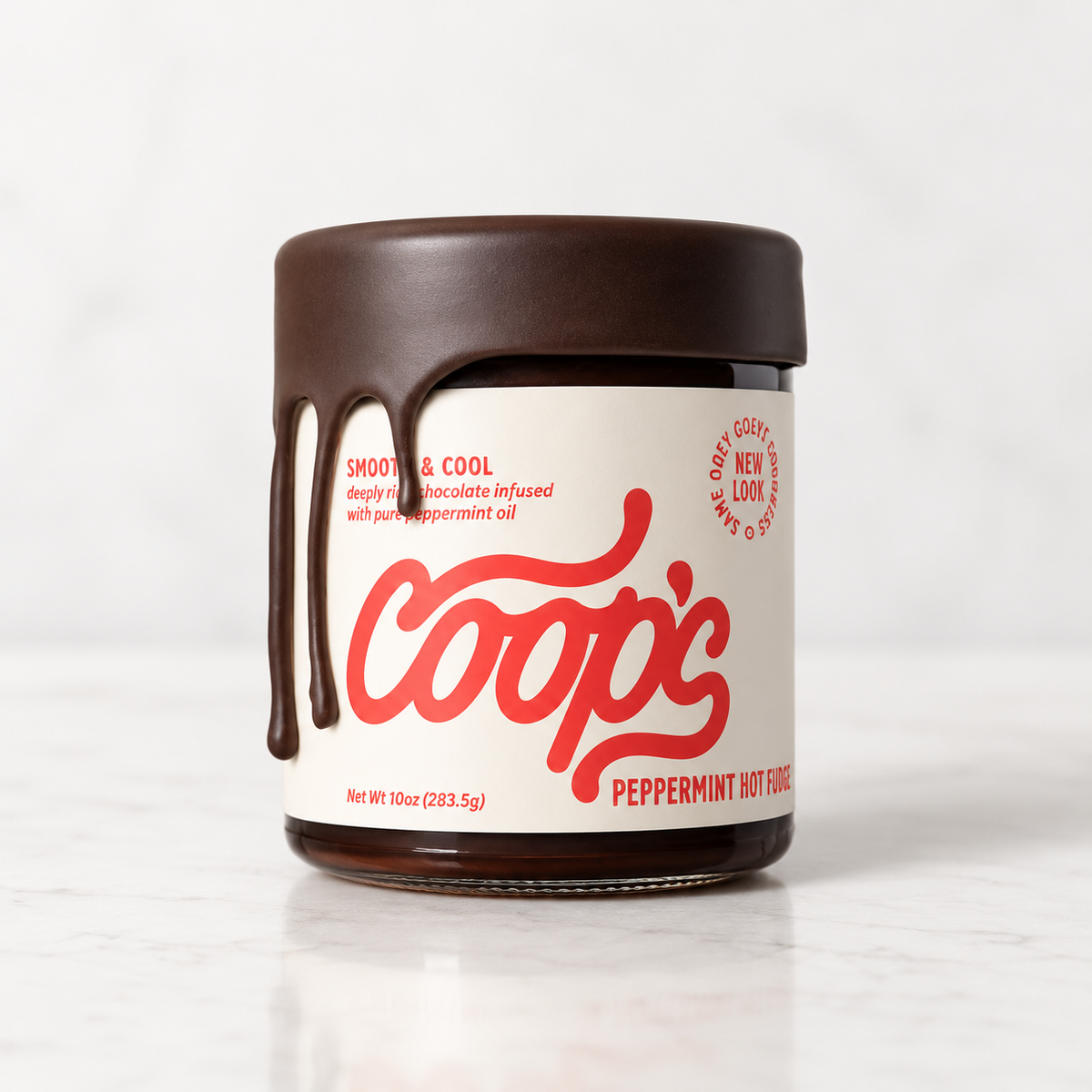 A jar of Coop's Peppermint Hot Fudge with a cream-colored label, red script logo, and dripping dark brown hot fudge on the side, set against a clean white marble background.