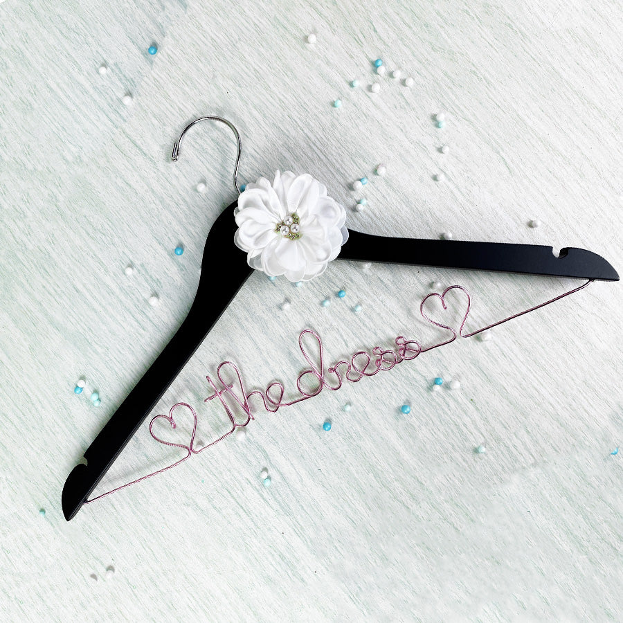 A black wooden clothes hanger is embellished with a white fabric flower and pink aluminum wire script that spells "The Dress," scattered with decorative beads.