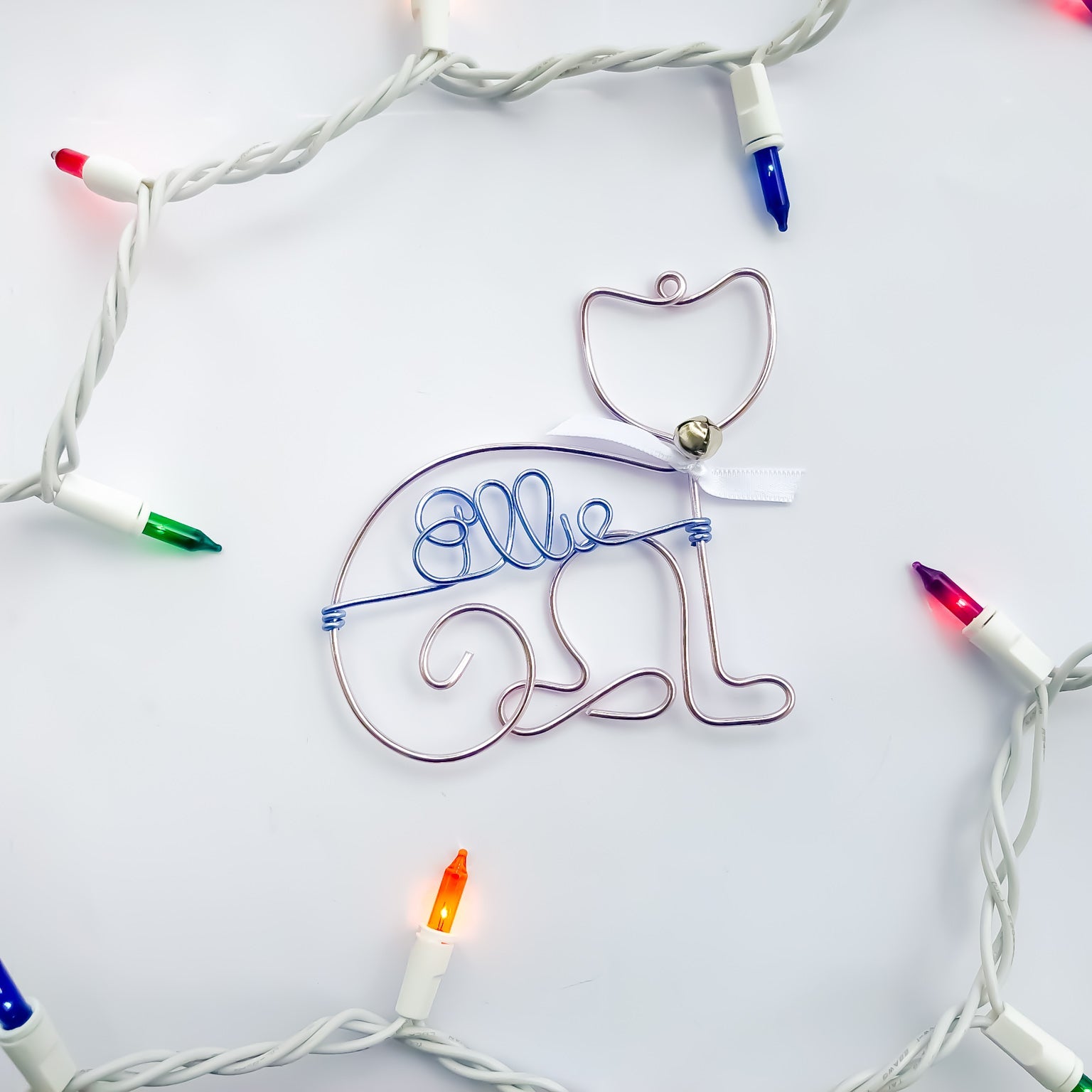 Personalized cat ornament, image shows the cat ornament done in rose gold wire that forms the outline of a cat's body and head with a white bow and a tiny silver bell. In the center of the ornament is personalized with ice blue colored wire with the name Ellie.