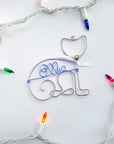 Personalized cat ornament, image shows the cat ornament done in rose gold wire that forms the outline of a cat's body and head with a white bow and a tiny silver bell. In the center of the ornament is personalized with ice blue colored wire with the name Ellie.
